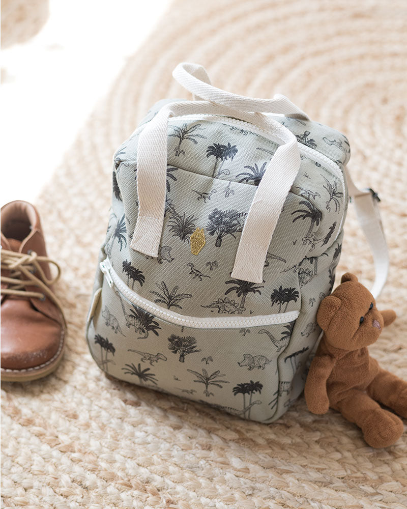 A Milinane toddler backpack with a dinosaur print, accompanied by a teddy bear and a pair of suede shoes, set on a textured circular rug.