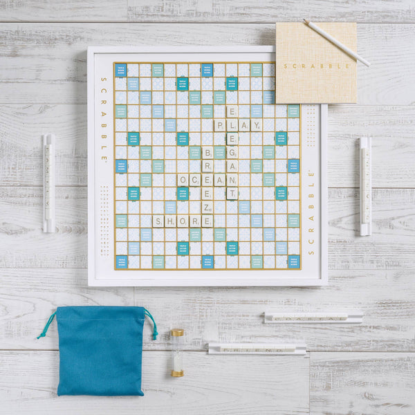 A Scrabble Bianco Edition game board set on a white surface with gold and blue grid squares, featuring letter tiles spelling “PLAY,” “OCEAN,” and “SHORE,” with four tile racks, a teal drawstring bag, a timer, and a score pad.