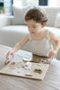 A young child with curly hair, dressed in light overalls, is focused on placing pieces into the Farm Animals Wooden Puzzle: A Tabitha Paige Puzzle on a table in a bright living room.