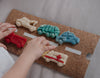 A child’s hands arrange colorful car-shaped dough pieces, made with the Service Vehicle Cutter Set - Eco Playdough Tool, on a corkboard with a painted white road—inviting creative sensory play.