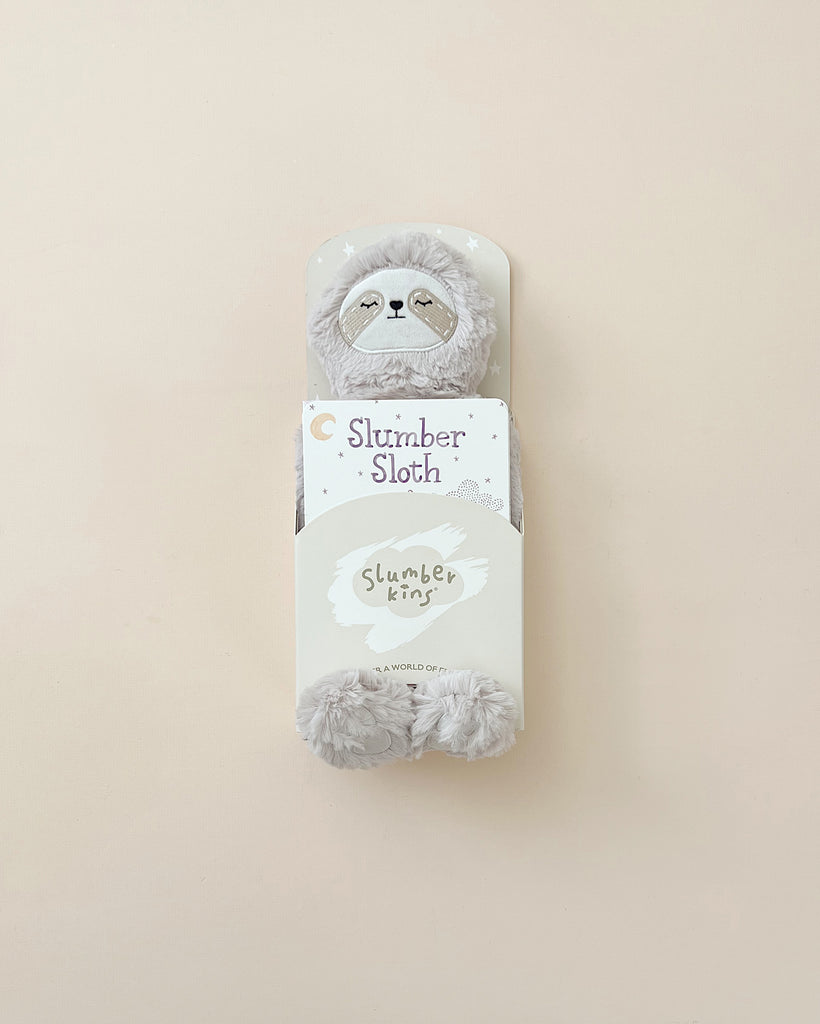 A pair of Slumberkins Sloth Kin slippers designed for children's routines, shaped like sloth faces, displayed in their packaging labeled "slumber sloth" on a light beige background.