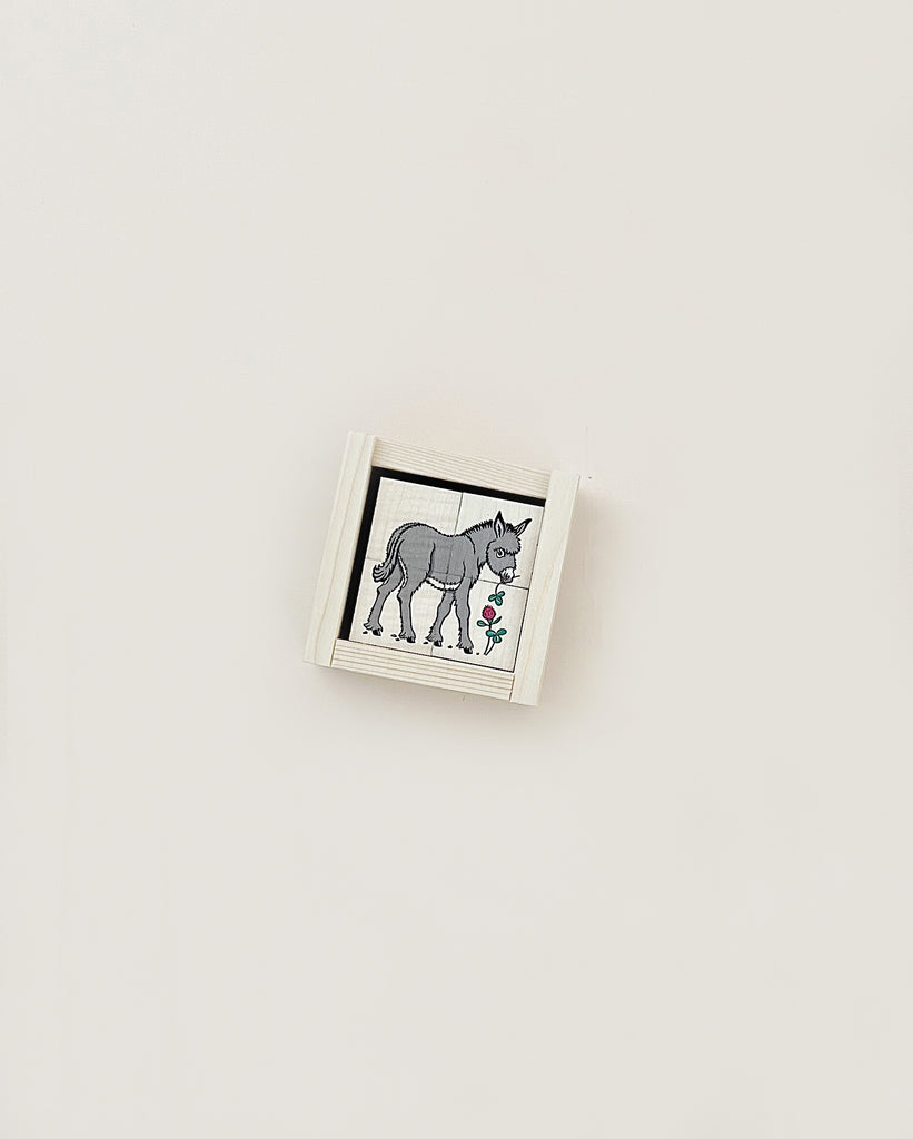A small framed illustration of a Wooden Block Puzzle - 4 Piece Farm Animals standing next to a flowering plant on a white background, crafted from sustainably harvested trees.