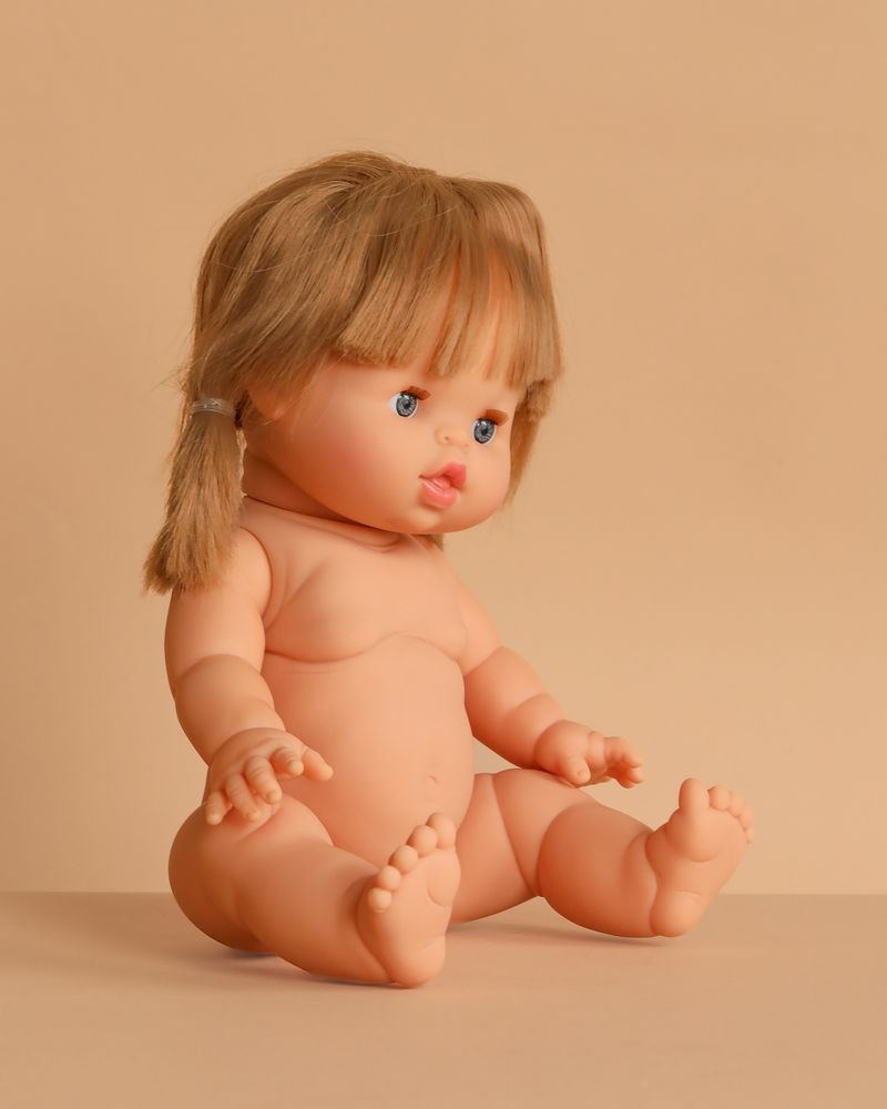 The Minikane Baby Doll (13") - Yzé sits upright on a beige surface with brown pigtails, blue eyes, and pink lips. Made in Spain, this anatomically correct doll comes unclothed; clothing sold separately.