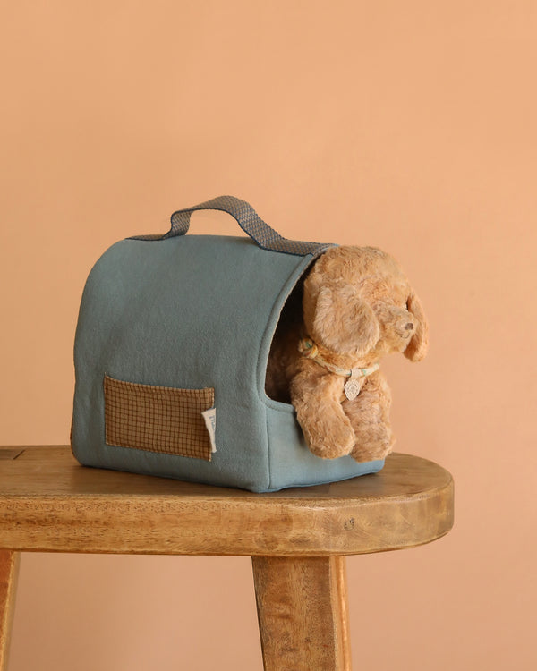 The Maileg Puppy in Dusty Blue Pet Carrier - Dusty Powder sits on a wooden stool against a beige background, ideal for displaying playful dog accessories.