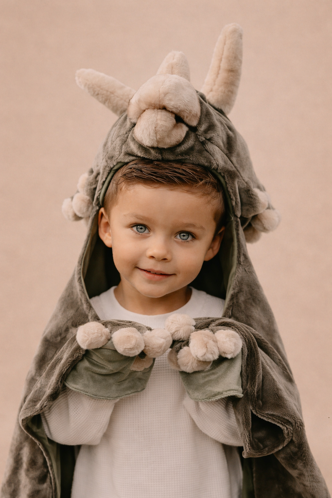 A young child with light eyes and short hair smiles in the Costume - Triceratops Dino, featuring horns and paw-like mittens, against a neutral background.