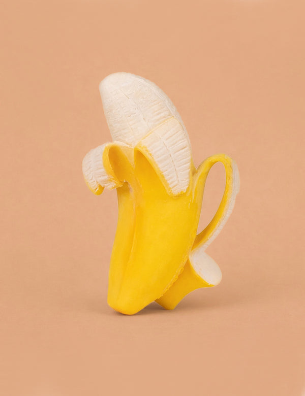 The Ana Banana Teether features a banana with its peel partially open, but the fruit is replaced by a white ceramic coffee cup on a solid light brown background, creating a whimsical sensory play toy.