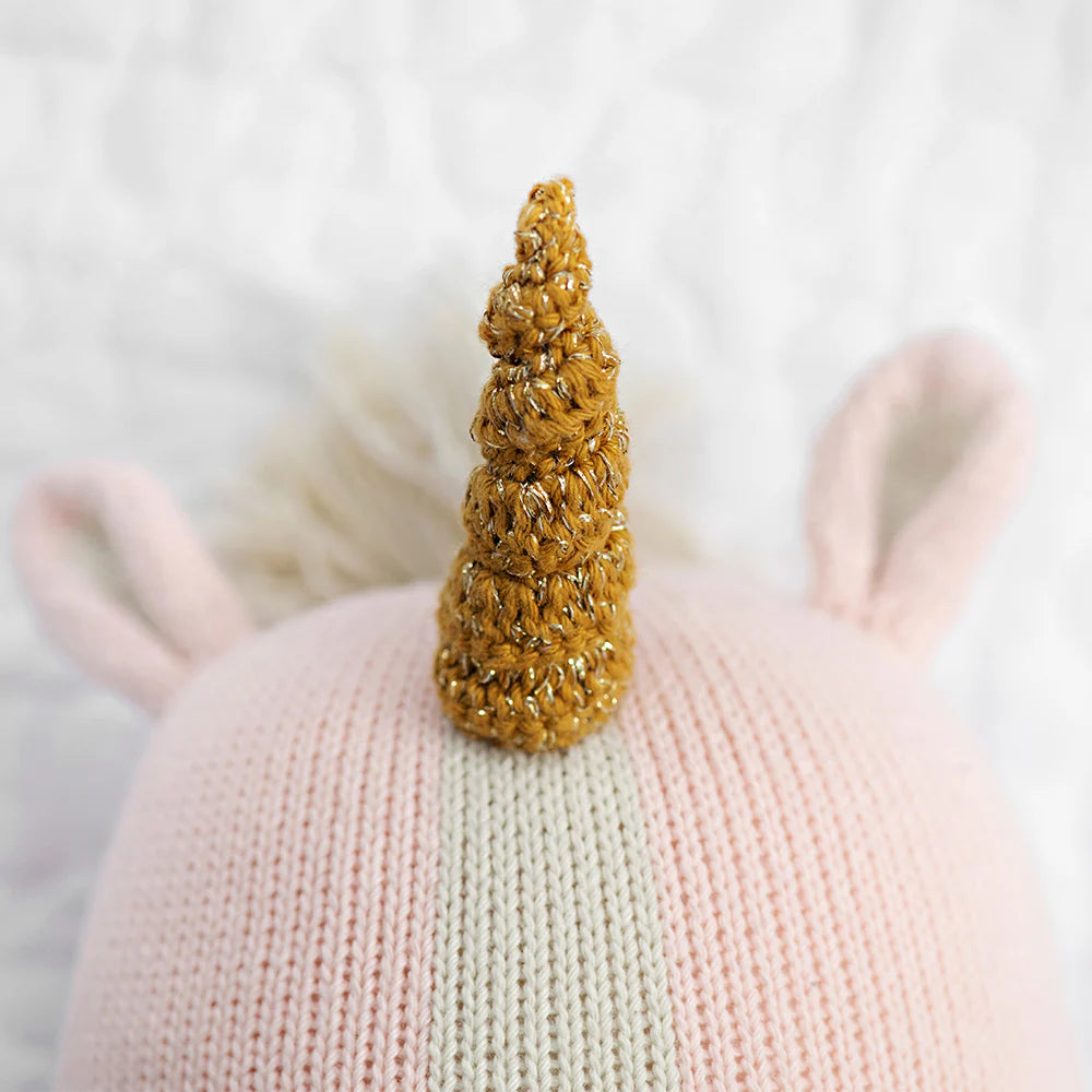 Close-up of a pink knitted hat with a gold crochet unicorn horn and light pink ears, designed as an accessory for the Cuddle + Kind Ella the Unicorn doll, shown against a white textured background.