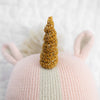 Close-up of a pink knitted hat with a gold crochet unicorn horn and light pink ears, designed as an accessory for the Cuddle + Kind Ella the Unicorn doll, shown against a white textured background.