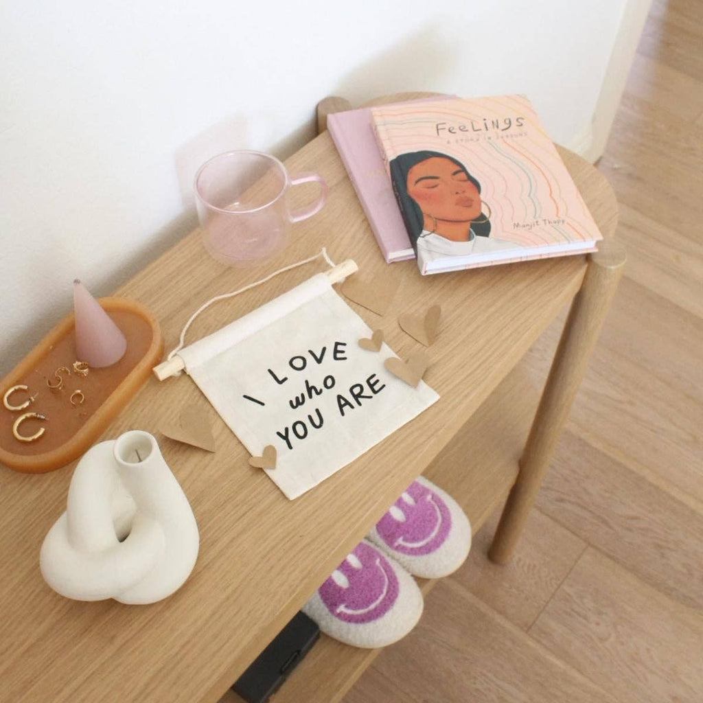A wooden table displays a handmade I Love Who You Are Canvas Sign - Natural, a pink cup, candle, jewelry dish, white vase, Feelings book, and smiley slippers—each item thoughtfully crafted by skilled Kenyan artisans.