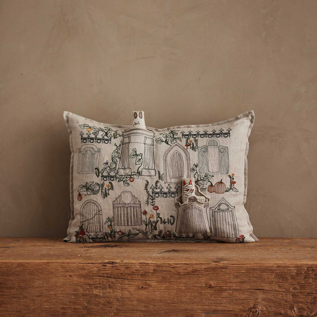 The Coral & Tusk Graveyard Pocket Pillow, featuring embroidered houses, plants, and flowers, is displayed on a wooden surface with two plush house ornaments and a small Bunny Ghost for spooky Halloween charm.