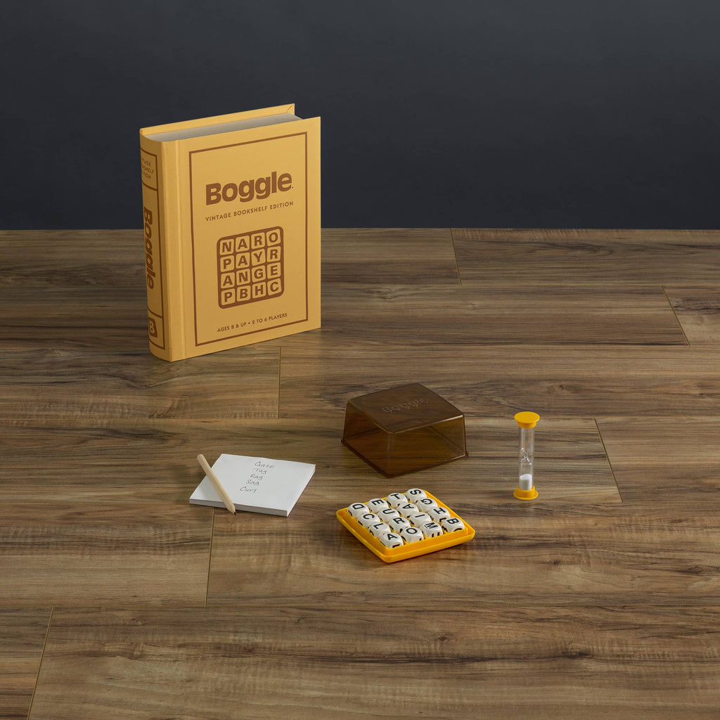 The Board Game: Boggle Vintage Bookshelf Edition is shown on a wooden surface, featuring a yellow box, lettered game grid, brown cover, hourglass timer, notepad, and pencil.