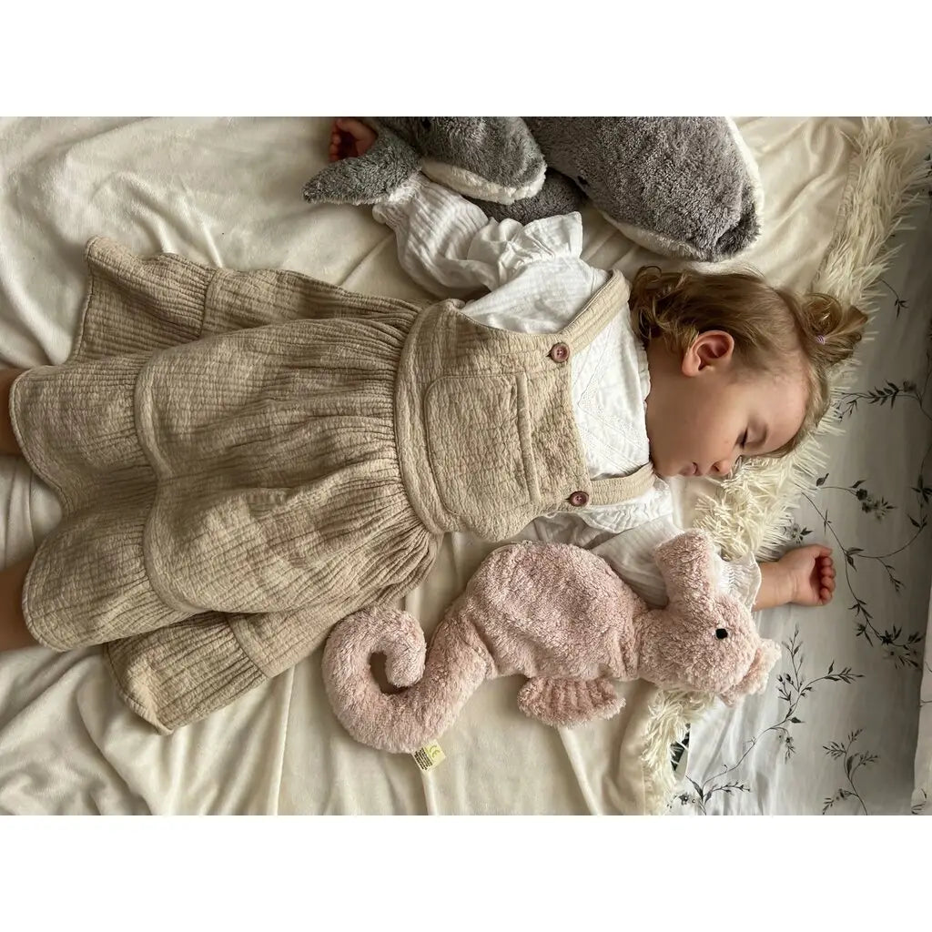 A toddler with light brown hair sleeps on a cream blanket, cuddling the Senger Naturwelt Cuddly Animal - Seahorse, with a pink seahorse grain pillow nearby. The child wears a beige pinafore dress over a white shirt.