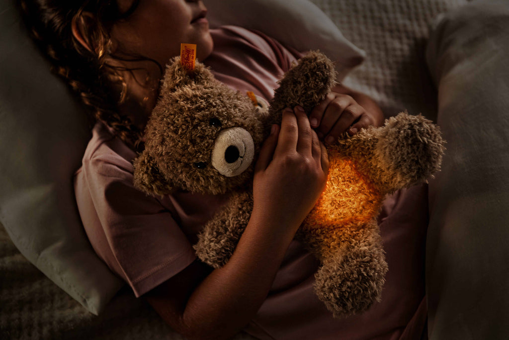 A child in bed cuddles a Light at Night Caspar Teddy Bear, whose softly glowing belly acts as a night light. Wearing a pink shirt and resting on a white pillow, the scene is cozy and warm.