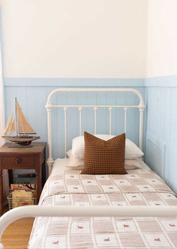A neatly made single bed is against a light blue paneled wall, topped with the Organic Cotton Twin Size Blanket - Pony. A brown checkered pillow features an embroidered pony, and a wooden nightstand holds a model sailboat and books.