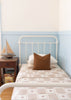 A neatly made single bed is against a light blue paneled wall, topped with the Organic Cotton Twin Size Blanket - Pony. A brown checkered pillow features an embroidered pony, and a wooden nightstand holds a model sailboat and books.