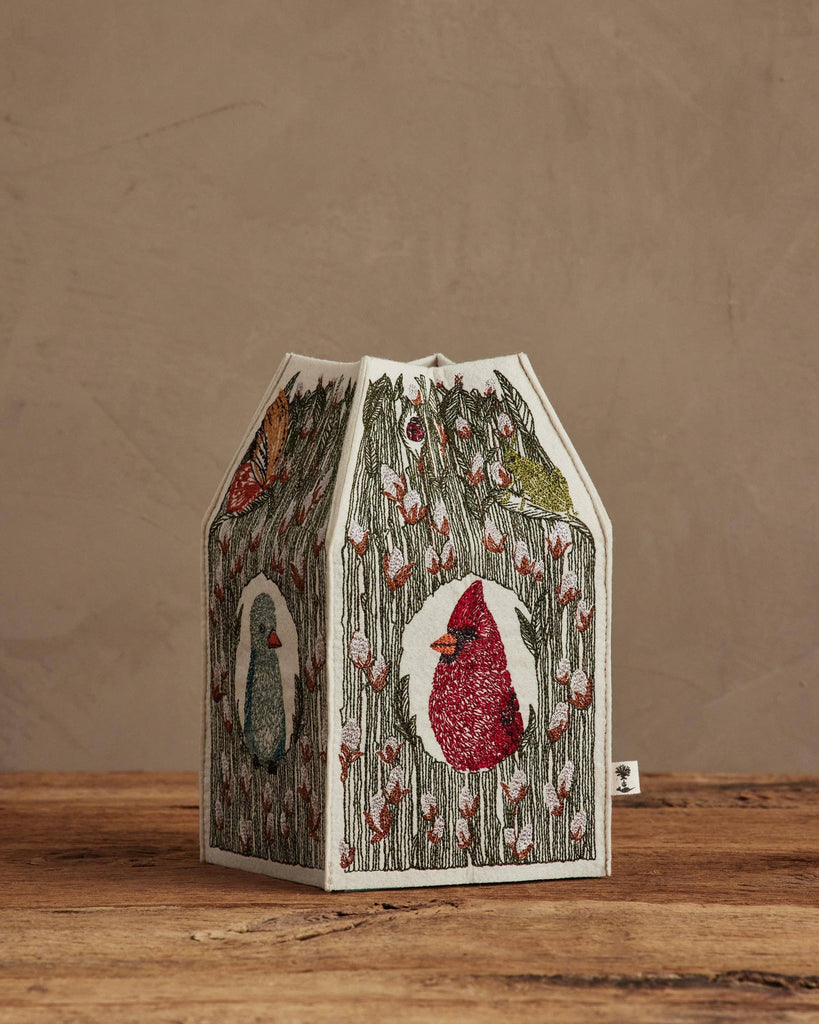 The Coral & Tusk Birdhouse Tissue Box Cover, featuring embroidered birds and floral designs, sits on a wooden surface against a beige wall.