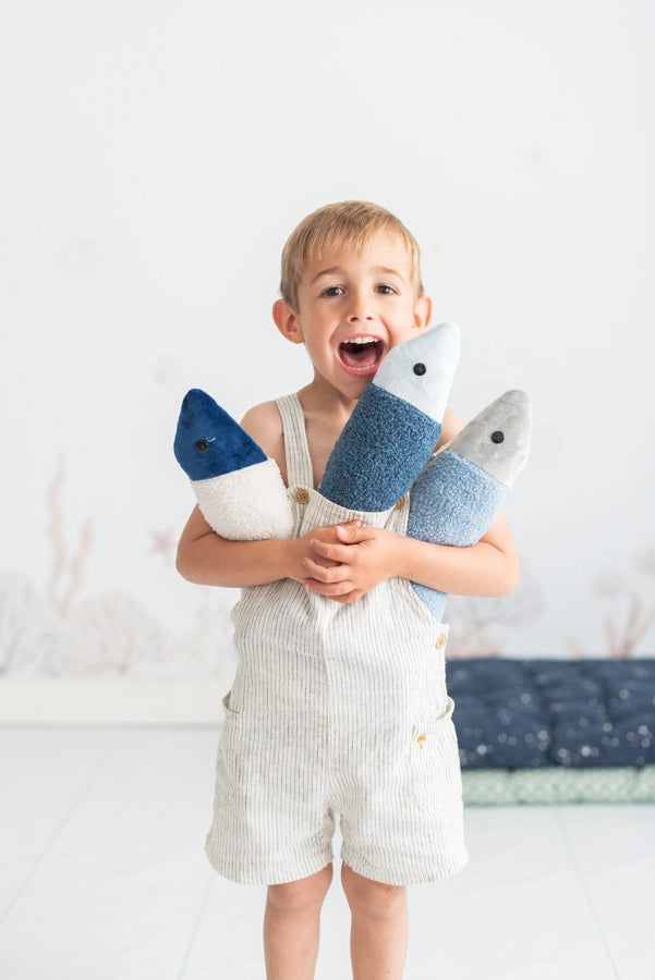A joyful young boy in a light-colored jumpsuit laughs as he holds two Dark Blue Fish Packs, one in each hand, made in Spain, in a bright, airy room with a cushioned background.