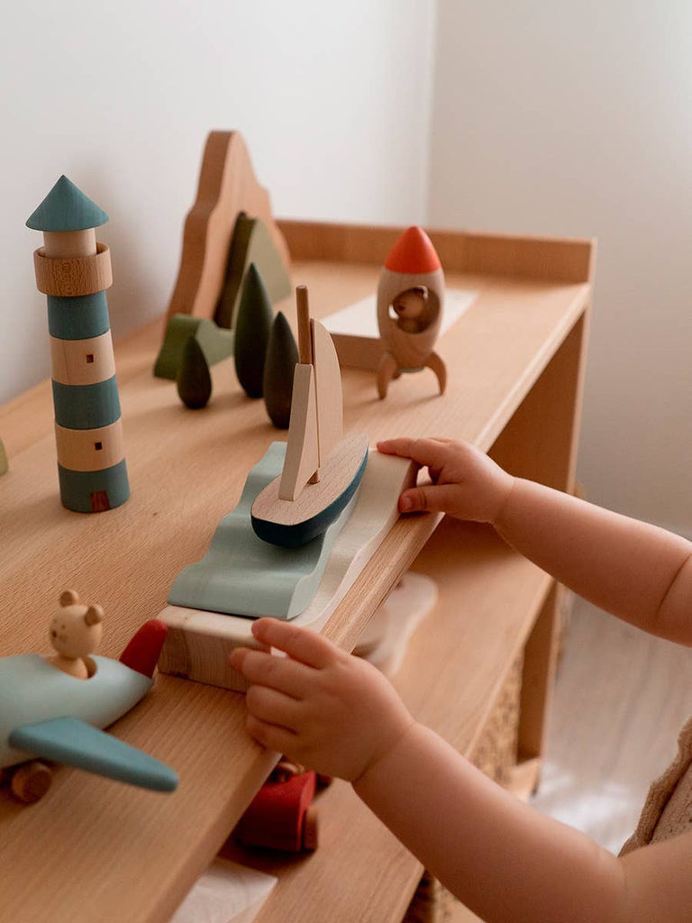 A child’s hands play with the Sabo Concept Sea Adventure Set on a shelf, featuring a lighthouse, sailboat, rocket, trees, and airplane in a softly lit and cozy scene.