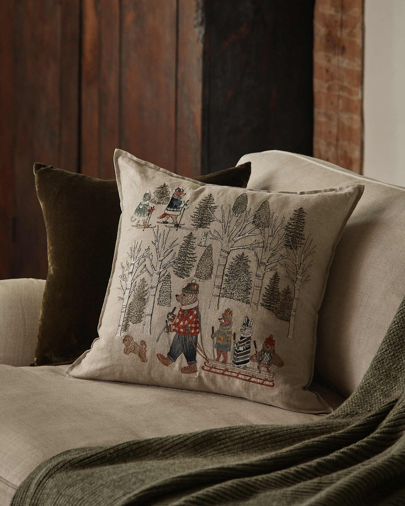 A beige couch features a green throw blanket and two pillows—one solid green and the Coral & Tusk Winter Fun Pillow, embroidered with woodland animals sledding and skiing—ideal for cozy winter or holiday decor.