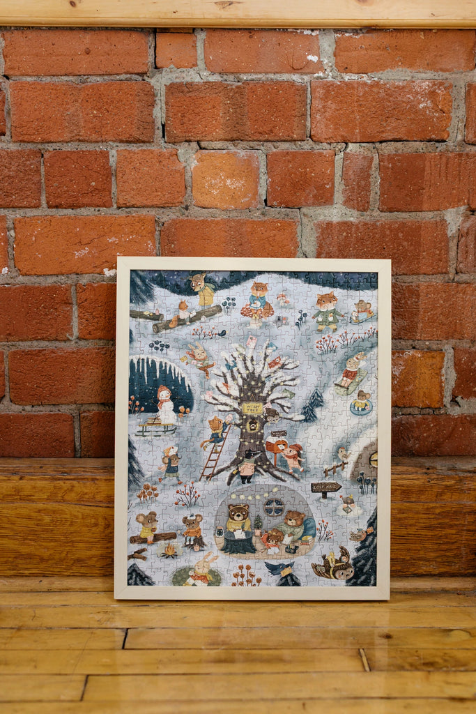 A completed Midnight Mail 500 Piece Puzzle, showing a whimsical winter scene with animals and people, is framed and leaning against a red brick wall on a wooden floor.