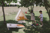 Two children play a regulation-sized Cornhole Game under the shade of trees on a grassy field. Nearby, a small tent is filled with yellow bedding and scattered wooden blocks. The scene is sunny and peaceful, ideal for outdoor play with all-weather bean bags ensuring the fun never stops.