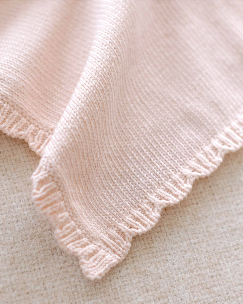 A close-up of the Cuddle + Kind Organic Cotton Baby Bunny Lovey (Blush), featuring soft, light pink hand-knit fabric with a scalloped edge and delicate ribbed border, displayed on a textured beige surface.