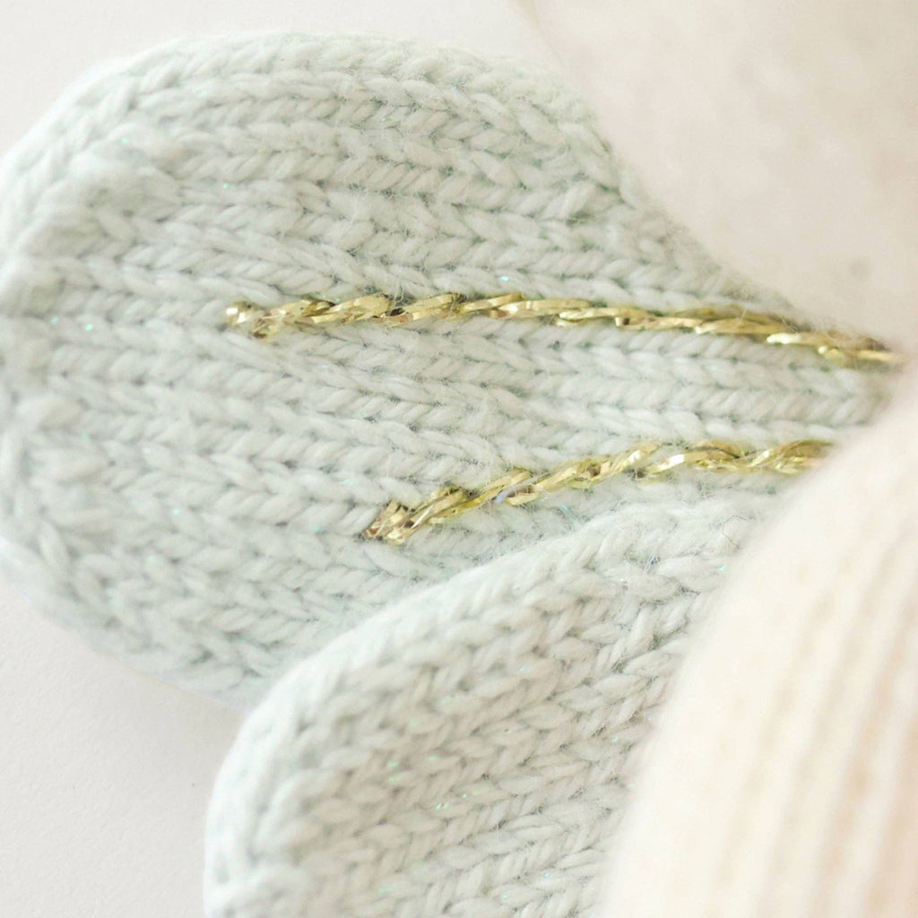 Close-up of a light blue knitted flower petal with shiny gold thread, from the handmade Cuddle + Kind Tiny Lily the Fairy doll, set against a soft white background.