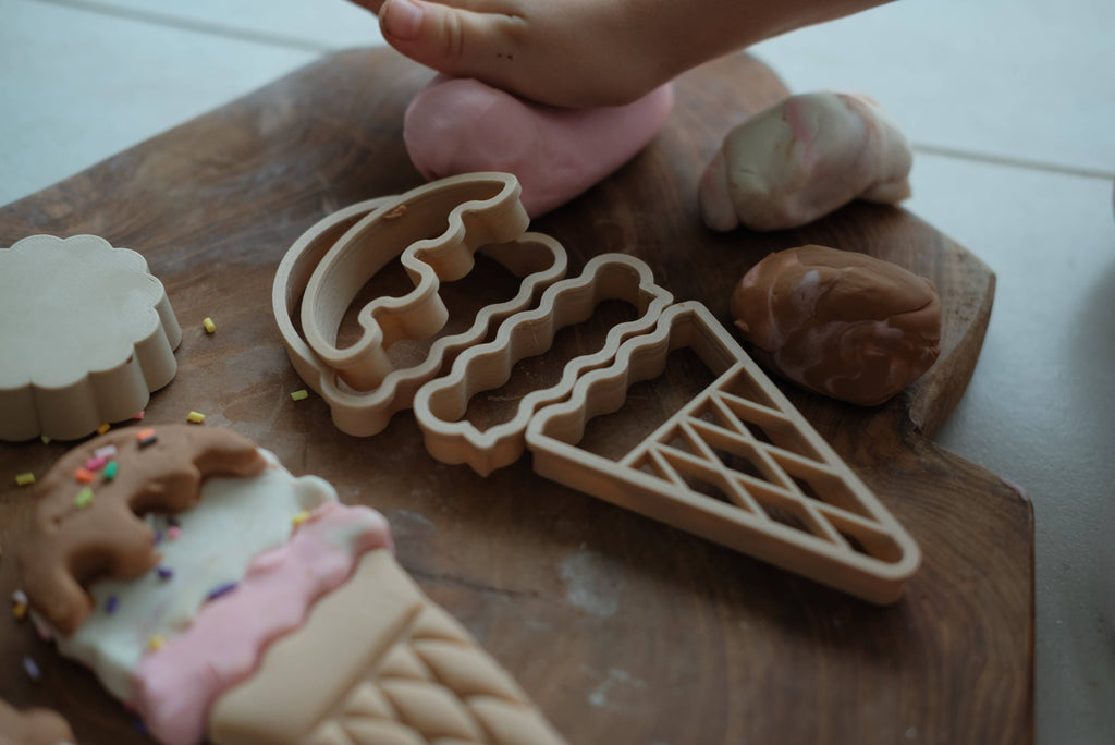 A child's hand presses pink dough next to the Ice Cream Cutter Set - Eco Playdough Tool, featuring wooden ice cream playdough cutters and pretend ice cream toys on a wooden board—perfect for imaginative sensory play.