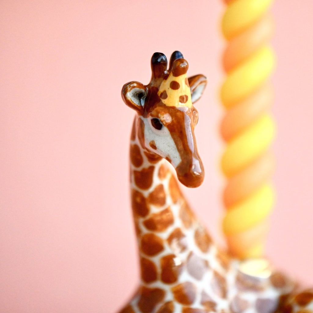 Close-up of the Giraffe Cake Topper, a fine porcelain figurine with detailed brown spots and tiny nubs on its head. The background features a peach color with a blurred yellow and white striped spiral, possibly a decorative candle, making it a charming collectible keepsake.