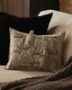 The Coral & Tusk Graveyard Pocket Pillow, featuring embroidered gates, a ghost, pumpkins, and flowers, sits on a beige sofa with a brown pillow and textured gray blanket.