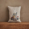 The Coral & Tusk Easter Bunny Pocket Pillow features an embroidered white rabbit in a scarf holding a basket with two smaller animals and colorful flowers. Displayed against a brown wall, it's perfect as an Easter decoration.