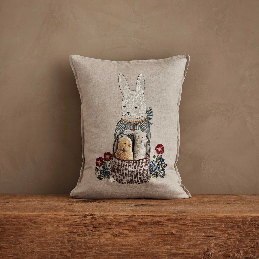 The Coral & Tusk Easter Bunny Pocket Pillow features an embroidered white rabbit in a scarf holding a basket with two smaller animals and colorful flowers. Displayed against a brown wall, it's perfect as an Easter decoration.