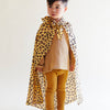 A young child stands against a plain background, wearing a tan outfit with ribbed leggings and a loose shirt. They have Sarah's Silks Animal Print Playsilk - Cheetah cape tied around their neck and look slightly to the side.