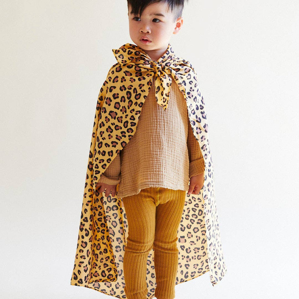 A young child stands against a plain background, wearing a tan outfit with ribbed leggings and a loose shirt. They have Sarah's Silks Animal Print Playsilk - Cheetah cape tied around their neck and look slightly to the side.