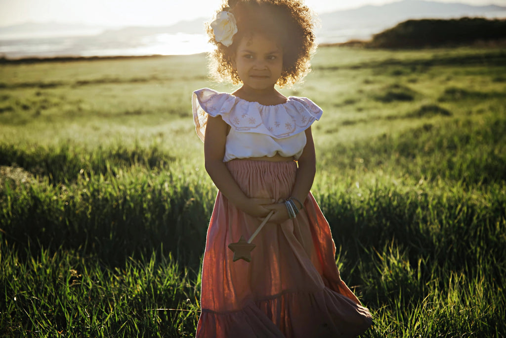 A young girl with curly hair standing in a sunlit field, dressed in a pink and white dress, holding a Wooden Star Wand from Bannor Toys, looking at the camera.