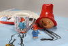 A ceramic, officially licensed Paddington Bear mug, a small figure, and the Paddington Money Bank with his blue coat and red hat are displayed on a themed cloth. Each item features Paddington in his red hat and blue coat.