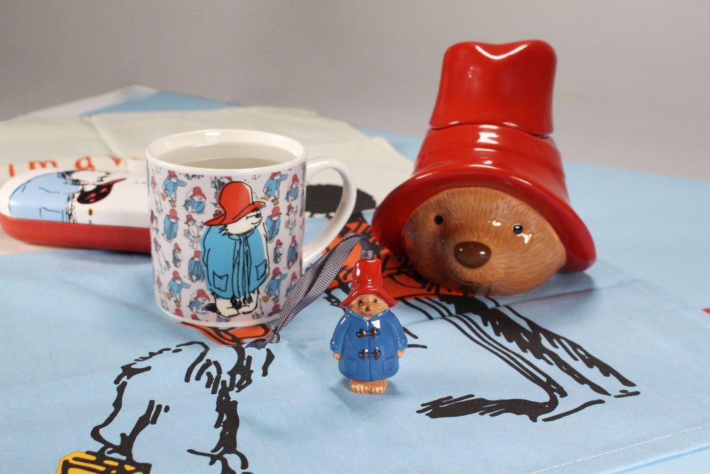 A ceramic, officially licensed Paddington Bear mug, a small figure, and the Paddington Money Bank with his blue coat and red hat are displayed on a themed cloth. Each item features Paddington in his red hat and blue coat.