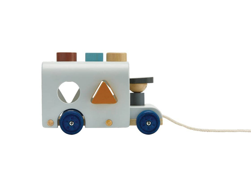 The Sorting Bus - Orchard Series is a wooden pull toy shaped like a vehicle, featuring blue wheels, colorful wooden pegs on top, and geometric shapes that fit into matching holes on the side for hands-on shape sorting fun. A string is attached to the front for easy pulling.