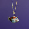 The Angler Fish Necklace features a handcrafted porcelain pendant shaped like a cartoonish anglerfish, complete with a wide grin, large eyes, and a small lure on top of its head. It hangs elegantly from a gold-plated chain against a solid purple background.