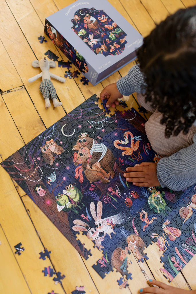 A child assembles the vibrant Courting Critters 500 Piece Puzzle on a wooden floor, with a stuffed toy and the puzzle box nearby—perfect inspiration for creating or gifting this charming, animal-themed puzzle for Valentine’s Day.