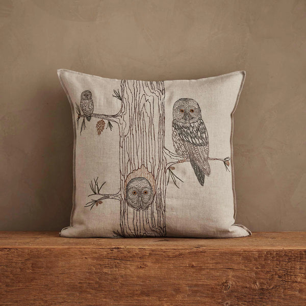 The Coral & Tusk Owl Family Tree Pillow is a decorative linen pillow featuring embroidered owls on tree branches, set against a neutral background and displayed on a wooden surface with a plain beige wall behind it.