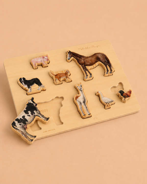A Tabitha Paige Farm Animals Wooden Puzzle showcases farm animal pieces—cow, pig, horse, sheep, goat, duck, and chicken—on a beige background. This charming educational toy is perfect for toddlers. In this scene, the cow piece is placed outside its slot.