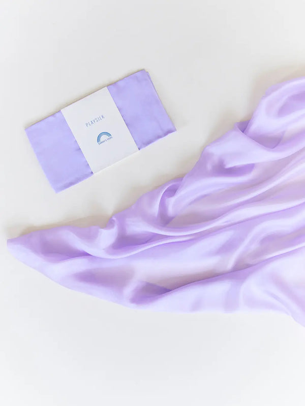 A Sarah's Silk Playsilk - Lavender in minimal packaging, placed next to a flowing, silky lilac playsilk on a white background. The ambiance is elegant and serene.