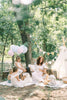 A woman reads Winnie-the-Pooh: Finding Feelings to six young children on a blanket in a sunlit forest, surrounded by balloons, bunting, and a small teepee for a festive touch.