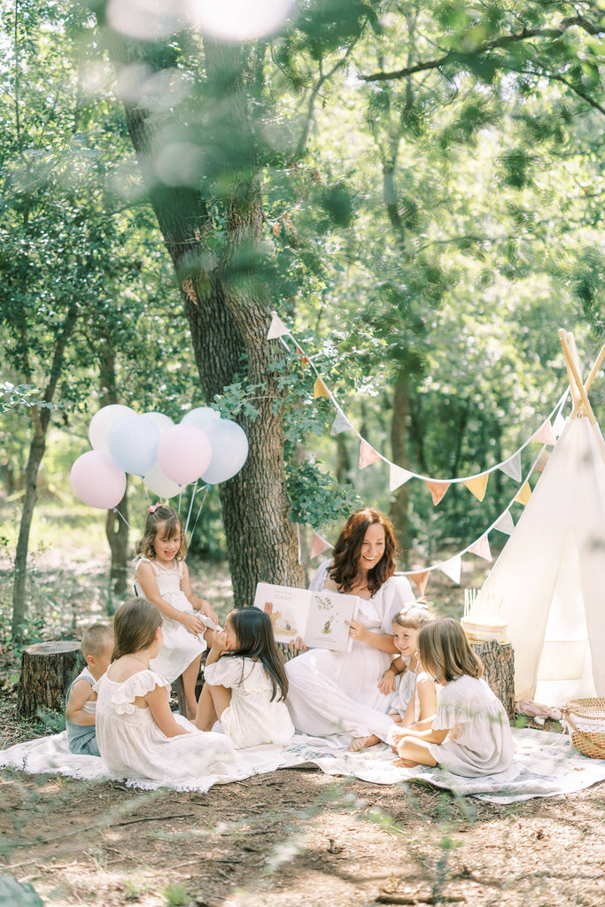 A woman reads Winnie-the-Pooh: Finding Feelings to six young children on a blanket in a sunlit forest, surrounded by balloons, bunting, and a small teepee for a festive touch.