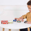 A young child in a mustard sweater plays with the London Toy Car Set—colorful, plastic-free wooden vehicles like a red double-decker bus—on a white table against a white background.