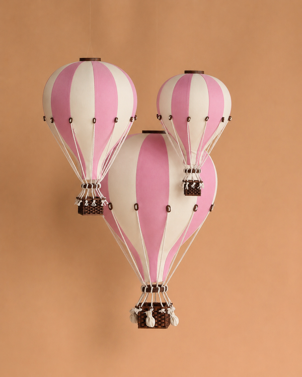 Three Hot Air Balloon - Powder Pink decorations with wooden baskets hang against a beige background, creating charming, handmade children's room decor. The powder pink balloons in various sizes appear to float in mid-air.