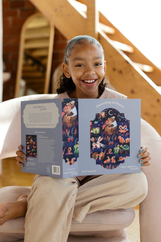 A smiling girl sits in a chair holding an open Courting Critters 500 Piece Puzzle box, which features colorful animal illustrations. Wooden stairs and a framed picture are visible in the background.
