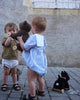 Two toddlers play outside on a cobblestone ground, developing their gross motor skills as they explore. One holds a teddy bear on wooden wheels, while the other faces away, holding another teddy bear. Nearby, Pull-Along Walter—a colorful pull-along toy dog—lies on the ground.