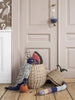 A whimsical scene featuring a Ferm Living Pear Braided Storage Basket - Large tipped over, spilling colorful stuffed toys, including a superhero and a bird, against a backdrop of a paneled wall with elegant bird wallpaper. The basket is crafted